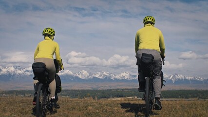 Naklejka na ściany i meble The man and woman travel on mixed terrain cycle touring with bikepacking. The two people journey with bicycle bags. Mountain snow capped.
