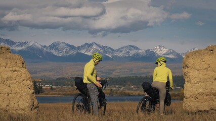 The man and woman travel on mixed terrain cycle touring with bikepacking. The two people journey with bicycle bags. Mountain snow capped, stone arch.