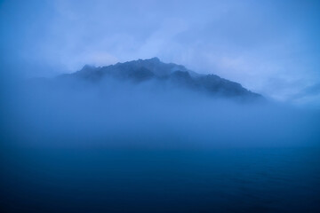 Big rocky mountain in middle of water among low clouds in dusk. Wavy sea of blue classic color. Atmospheric minimalist landscape with rock on lake in dense fog. Misty scenery with deep blue calm water