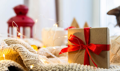 Gift with a red ribbon on a white knitted blanket. New Year, garlands, gifts, a cup and marshmallows - an atmosphere of warmth, comfort and magic.