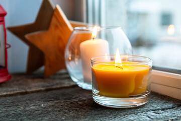 Cozy atmosphere - candles on a wooden window sill at dusk. Candles in glass candlesticks and wooden stars in the background.