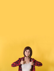 Portrait beautiful young woman standing in checked shirt and gave thumbs up: vertical banner with space for text, well signed sign and looking at the camera on yellow background