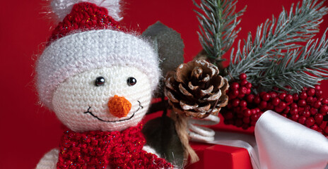 Snowman toy in a Santa Claus hat on a red background next to a red gift box tied with a white ribbon and an evergreen spruce twig with frost, cones and berries, crop banner
