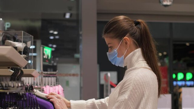 Skilled designer in blue disposable mask looks at different coloured cotton pullovers combining new winter outfit. Concept style and design