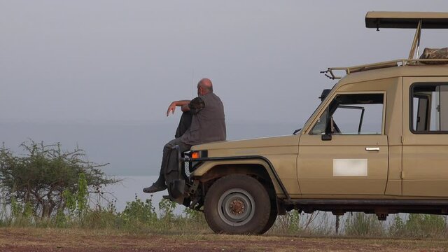 A Male Tourist Watches Africa Landscape by Safari Vehicle. Watches sitting journey travel holiday voyage visit solo single alone look looking old man people Toyota Land Cruiser see trip tour hairless.
