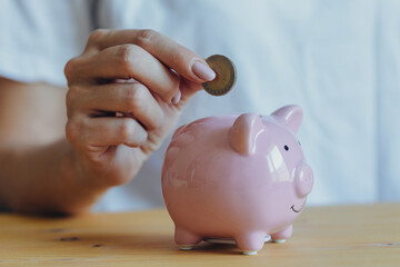 the woman's hand puts a coin in a pink piggy bank standing on the table. Concept of saving money or savings, investment during the global crisis