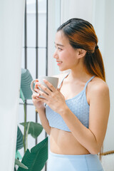 Beautiful sporty asian woman holding cup of coffee looking outside the window.