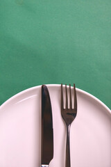 Vintage silverware on dark green background. White plate with knife and fork over green table, flat lay.