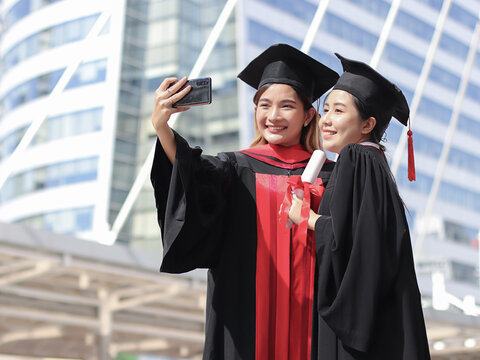  happy graduated women in graduation gowns making selfie photo with mobile phone. City building background. Education, Friendship and technology concept..