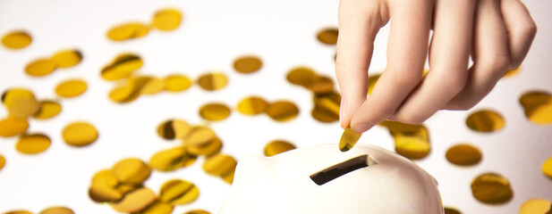 Hand putting coin to piggy bank. White piggy bank on background of golden paper confetti isolated on white background. Saving money concept