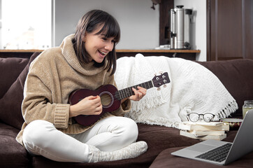 Young beautiful woman learning to play ukulele at home with online lessons.