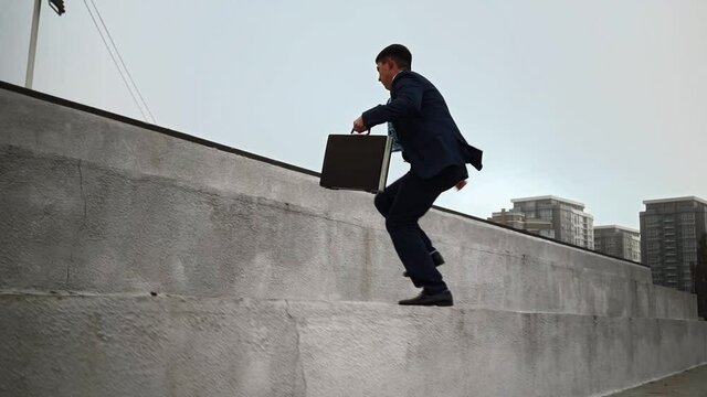 A young businessman in a suit with a suitcase in his hands jumps up the parapets against the city background. Career stairs concept. On the way to success. Corporate promotion. Slow motion.