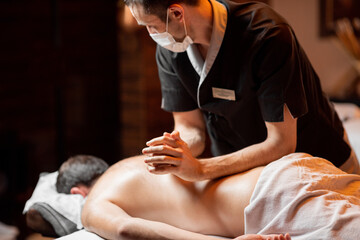 Professional masseur in facial mask doing a deep massage to a male client at Spa salon. Business during the epidemic concept