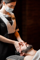 Professional female masseur in medical mask doing facial massage to a male client at Spa salon. Business during the epidemic concept