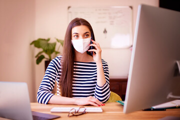 Young woman talking with somebody on her mobile phone while using computer and working from home office