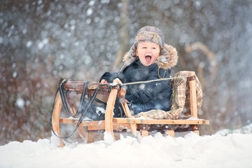 a boy in an overalls with fur and a hat with earflaps sits in a wooden sled. snowing winter. child...