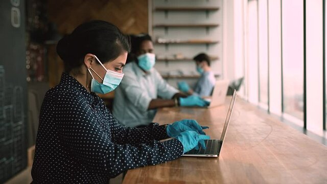 Indian office employee and colleagues multi ethnic company staff wear surgical facial masks and protective gloves working on laptops sit in shared table in coworking, keeping distance due coronavirus