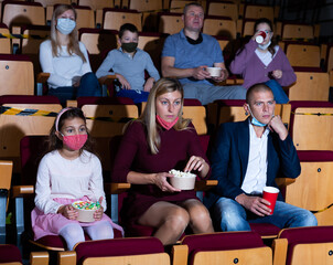 Family with child wearing protective masks eating popcorn and watching a movie in the cinema