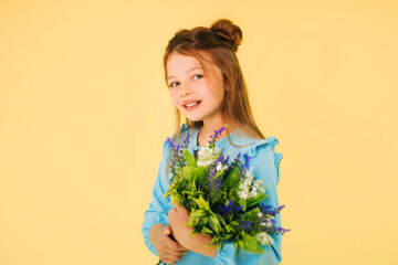 A girl of twelve years old in a blue dress on a yellow background holds lavender and lily of the valley flowers in her hands. Fashionable child. Studio photo