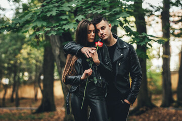 Gorgeous fashionable couple in black posing outside with red rose in hands, looking unemotional and...