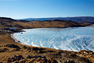 View of frozen Lake Baikal in winter. Small Sea Strait in sunny frosty day, transparent smooth ice with cracks. Tourist wooden hotel houses  on the lake, tourists walk along the frozen Bay.