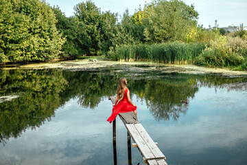 Fototapeta na wymiar Back view full length portrait of young pretty woman sitting on the wooden pier near the river or lake touching clothes, dressed in red dress, looking away, copy space and nature blur background.