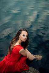 Vertical portrait of young pretty wet woman mermaid sitting on pier rock in the water river or lake, dressed in red dress, looking at the camera, copy space and nature blur background.
