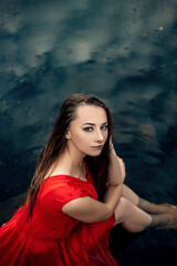 Vertical portrait of young pretty wet woman mermaid sitting on pier rock in the water river or lake touching head, dressed in red dress, looking at the camera, copy space and nature blur background.