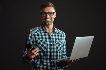 Smiling unshaven man in eyeglasses holding laptop and cellphone