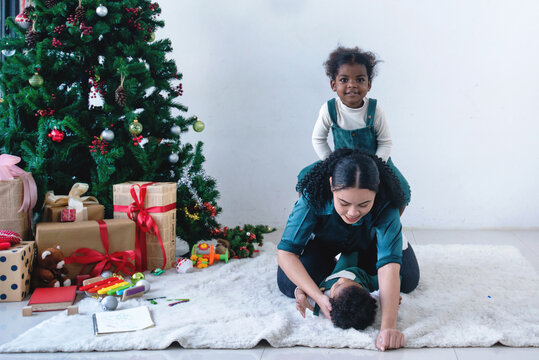 Dark Skinned Daughter Enjoyed Climbing On Her Mother's Back., Mom Is Teasing Her Son On The Floor, Near Christmas Tree