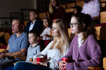Fototapeta premium Friendly family of four sitting with popcorn and drinks in cinema, watching movie with interest. Family leisure and entertainment concept