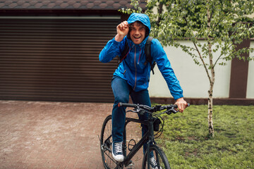 Happy curly male courier in blue raincoat delivers parcel cycling with a bicycle. Joyful young man standing with his bike before bicycling on a rainy day next to the house.