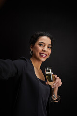 Vertical portrait of elegant Middle-Eastern woman holding champagne glass and taking selfie photo while standing against black background at party, copy space © Seventyfour