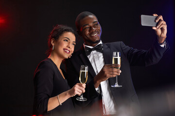 Waist up portrait of elegant mixed-race couple taking selfie photo while standing against black background at party, copy space