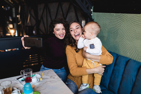 Two friends enjoying in cafe bar with cute little baby boy and taking selfie photo.