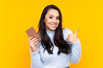 Young Colombian girl over isolated yellow background taking a chocolate tablet and with thumb up