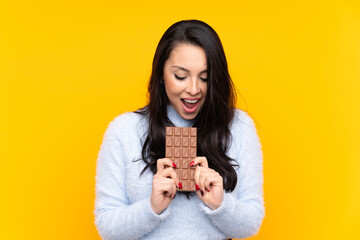 Young Colombian girl over isolated yellow background eating a chocolate tablet