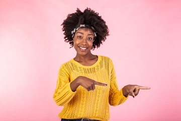 Horizontal shot pleased dark skinned female with Afro haircut, points right with both fore fingers, shows blank space for your promotion, isolated over pink background. People, advertisement concept