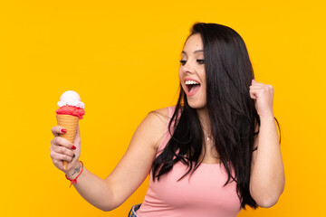 Young Colombian girl holding an cornet ice cream over isolated yellow background celebrating a...