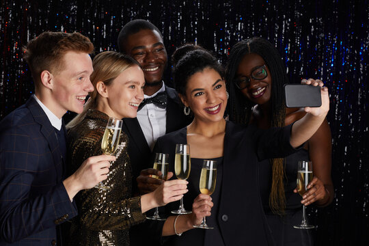 Waist up portrait of multi-ethnic group of friends holding champagne glasses and and taking selfie together while enjoying elegant party