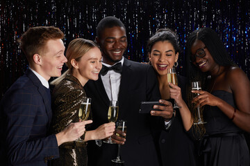 Waist up portrait of multi-ethnic group of friends holding champagne glasses and livestreaming online while enjoying elegant party