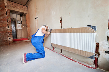 Plumber in work overalls using wrench while installing heating radiator in room. Man in helmet installing heating system in apartment. Concept of radiator installation, plumbing works, home renovation