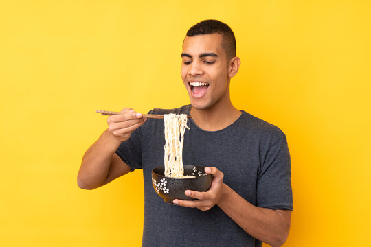 Young African American man over isolated yellow background holding a bowl of noodles with chopsticks and eating it