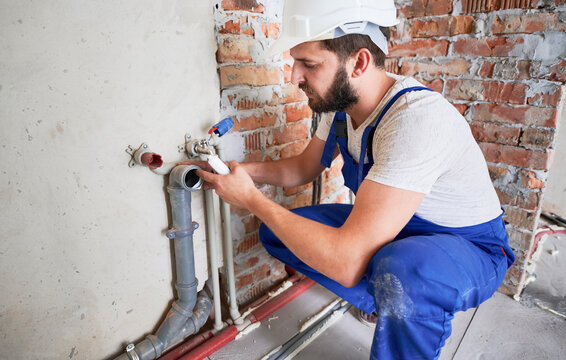 Young plumber, wearing blue uniform and white helmet working with sealant fix of sewer pipe in kitchen or in bathroom in unfinished apartment. Concept of plumbing works and home renovation