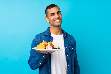 Young handsome man holding waffles over isolated blue background with happy expression