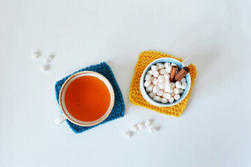 Mustard yellow and dark blue handmade knitted coasters, cups with tea, marshmallows and cinnamon over white