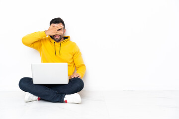 Caucasian man sitting on the floor with his laptop covering eyes by hands and smiling