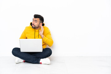 Caucasian man sitting on the floor with his laptop listening to something by putting hand on the ear
