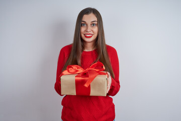 Fototapeta premium Beautiful surprised woman in a red sweater, holding a gift box, looking at the camera on a gray background. The concept of holiday, Christmas, New Year