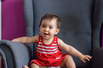 Niña pequeña simpática y sonriente con vestido rojo con lineas blancas sentada en sillón gris
